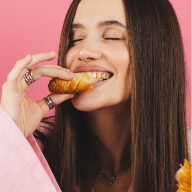 Donna sorridente gusta un croissant con piacere, sfondo rosa vivace.